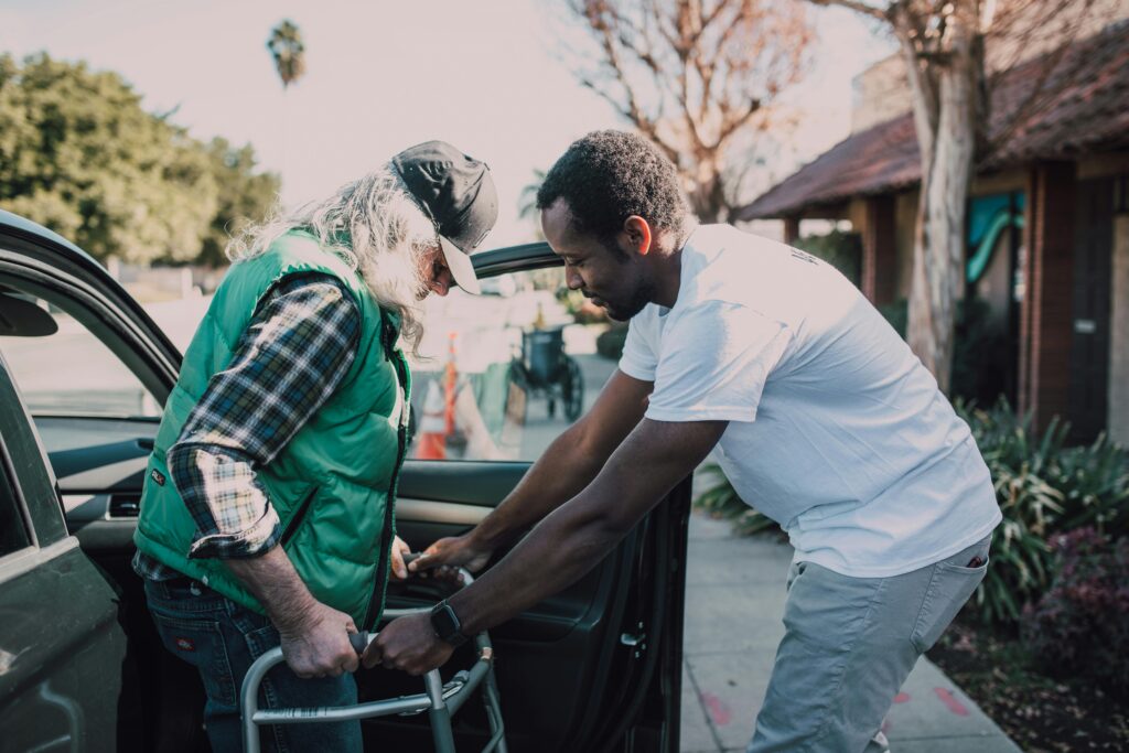 pexels-photo-6647037-6647037 Young volunteer helping senior man with walker near a parked car, symbolizing care and support.